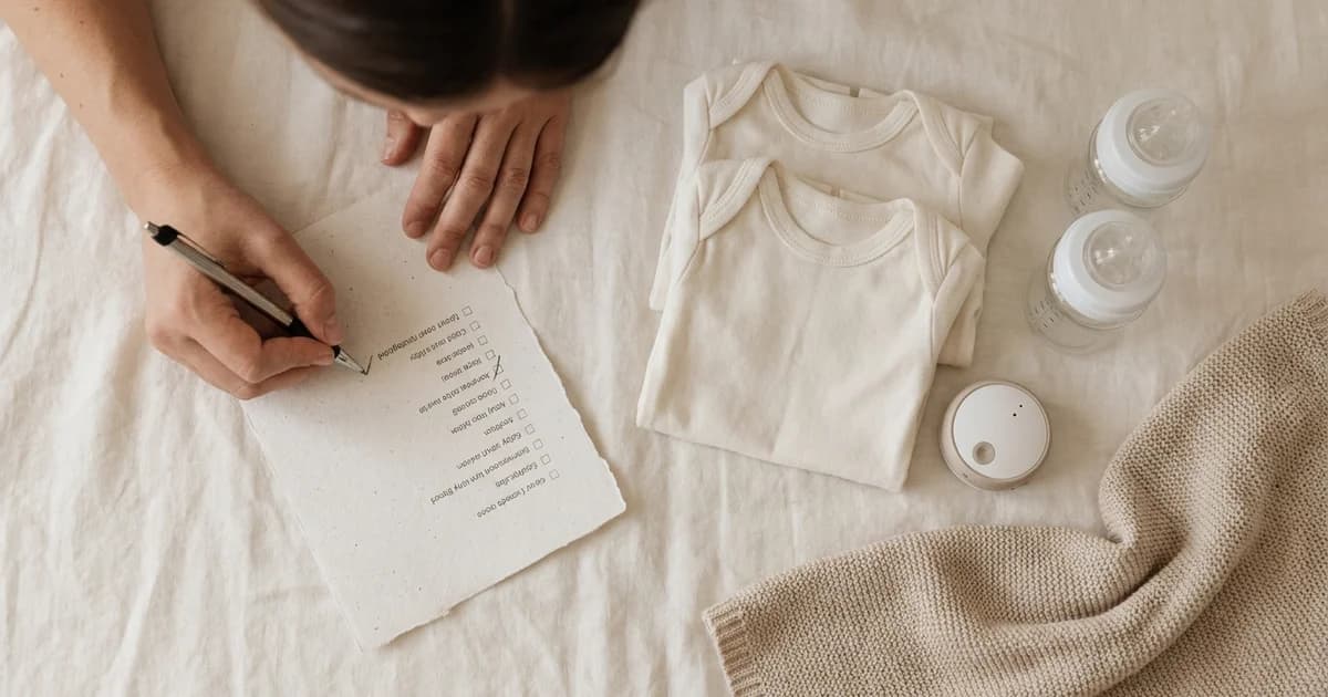 Parent checking items off a baby registry checklist beside organized baby gear - onesies, bottles, and a soft blanket
